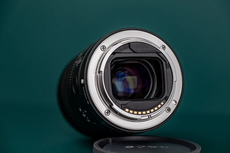 A Sony E-mount camera lens is positioned on a dark green surface, showing its rear metal mount, electronic contact pins, and internal glass elements, with a lens cap lying in the foreground.