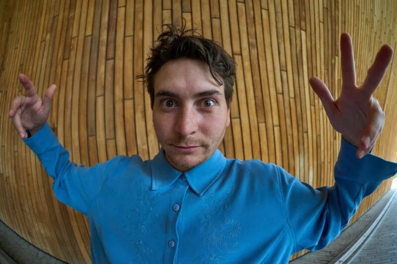 A person with messy brown hair wearing a blue embroidered button-down shirt poses with both hands raised in front of a curved wooden wall.