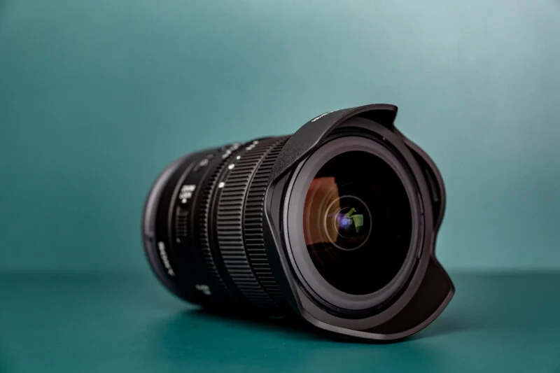 A black camera lens with a petal-shaped hood rests on a solid teal surface against a matching teal background.