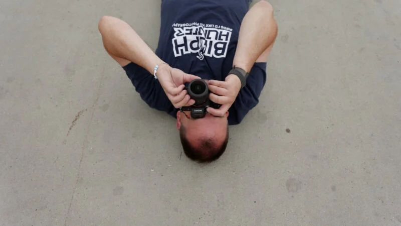 A person wearing a blue t-shirt with the text "BIRD HUNTER" lies on their back on a concrete surface, holding a camera up to their face.