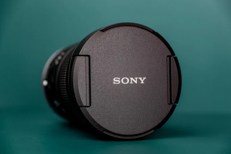 A black Sony camera lens cap sits on a teal surface, angled slightly to show the side of the lens barrel.