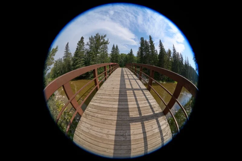 A wooden footbridge with brown railings stretches over a shallow stream, framed by a circular fisheye lens effect.