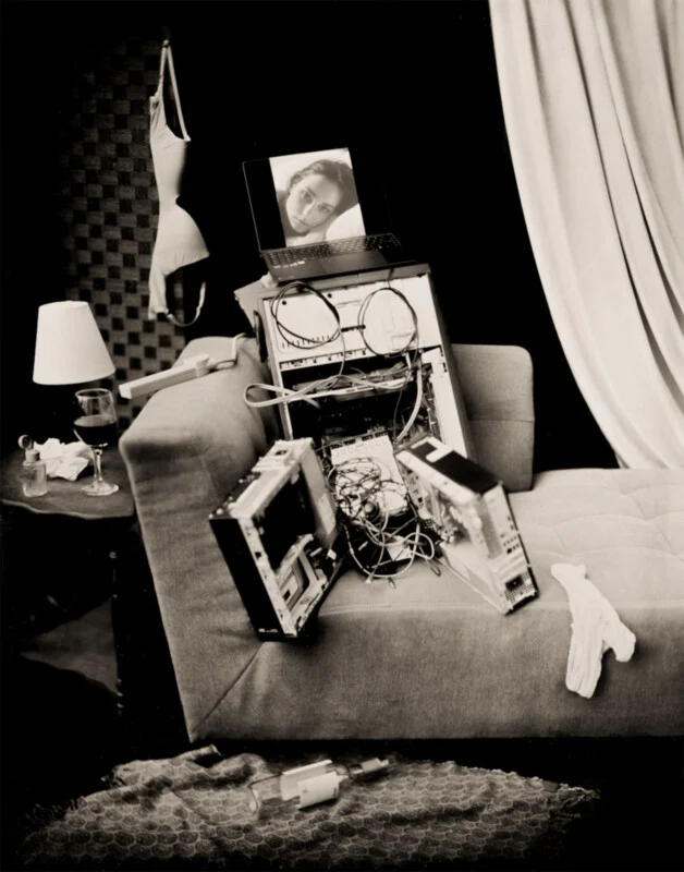 A disassembled computer tower sits on a sofa, topped with a laptop displaying a woman's face, near a side table with a lamp, wine glass, and hanging lingerie.