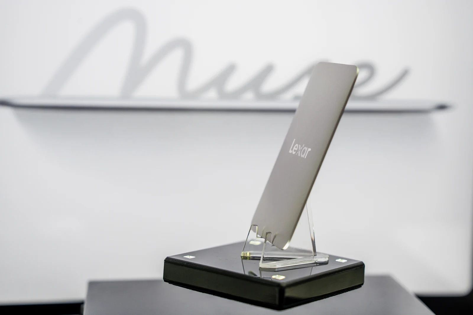 A slim, silver Lexar portable solid-state drive stands upright on a clear plastic display stand atop a black base, with the word "Muse" written in cursive on the wall behind it.