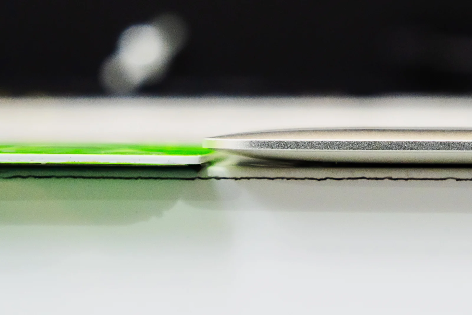 A side-by-side close-up shows a thin, flat green card next to a thicker, rounded metallic object resting on a white surface.
