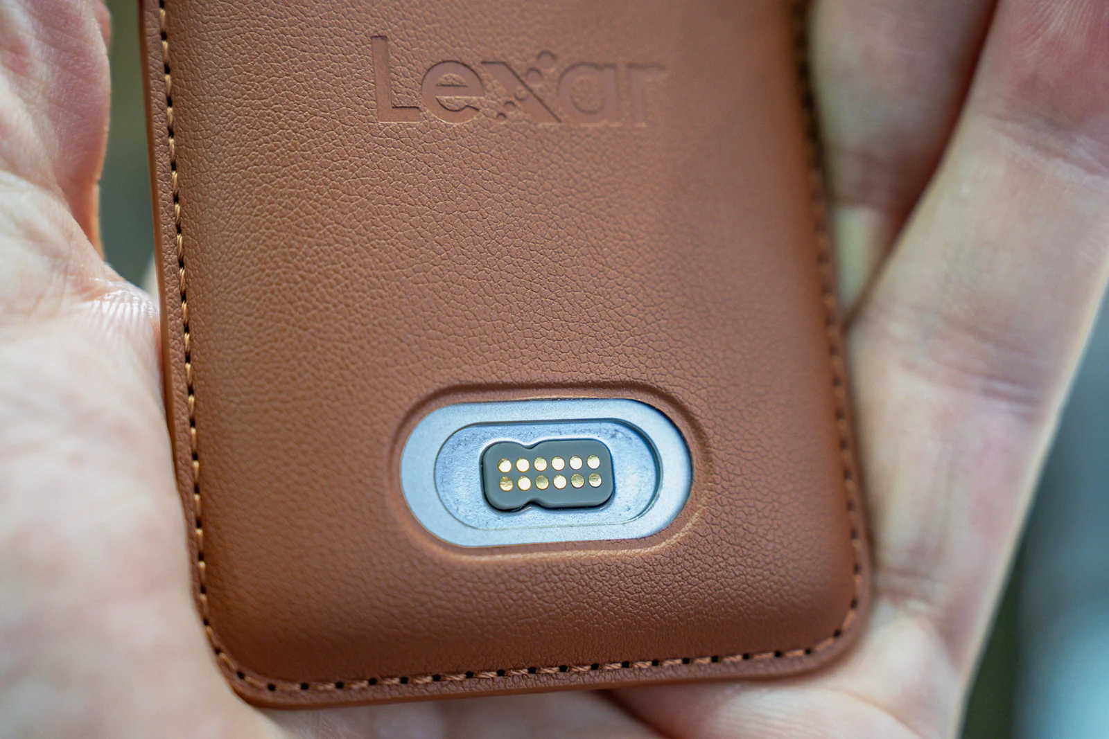 A hand holds a brown leather-textured device with the brand name "Lexar" embossed at the top and a metallic, ten-pin charging contact port centered below.
