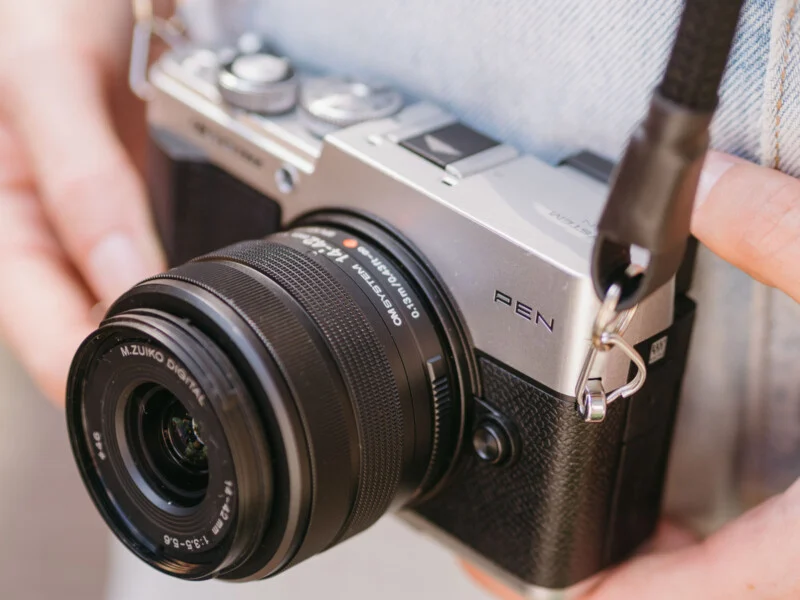 A person holds a silver and black OM System PEN digital camera with an M.Zuiko Digital 14-42mm lens attached.