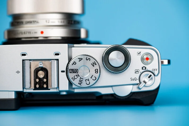 A top-down view of a silver digital camera body with a lens, featuring a mode dial, shutter button, and power switch against a blue background.