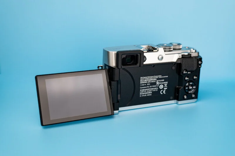 A silver and black digital camera with its rear LCD screen flipped out to the left, set against a solid light blue background.