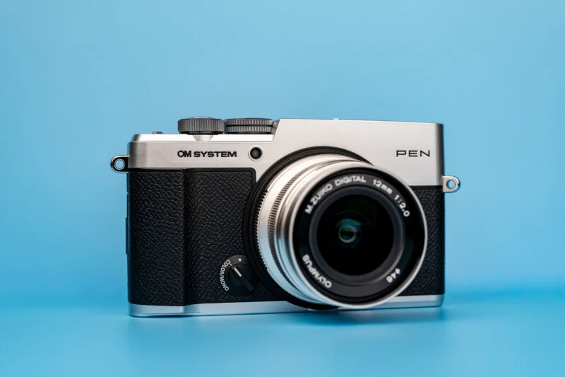 A silver and black OM SYSTEM PEN digital camera with a lens attached, centered against a solid light blue background.