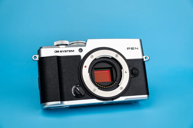 A silver and black OM SYSTEM PEN mirrorless camera body with an exposed sensor, set against a solid blue background.