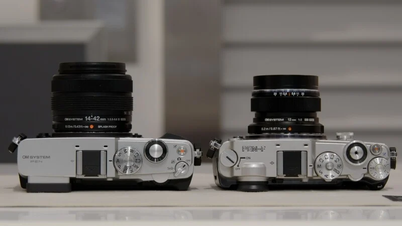 Two silver mirrorless cameras sit side-by-side on a white surface, each equipped with a black lens.