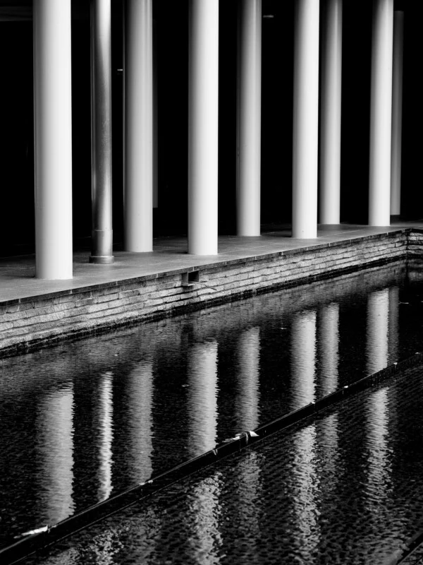 A row of white cylindrical columns stands beside a dark reflecting pool, with the pillars mirrored in the rippling water.