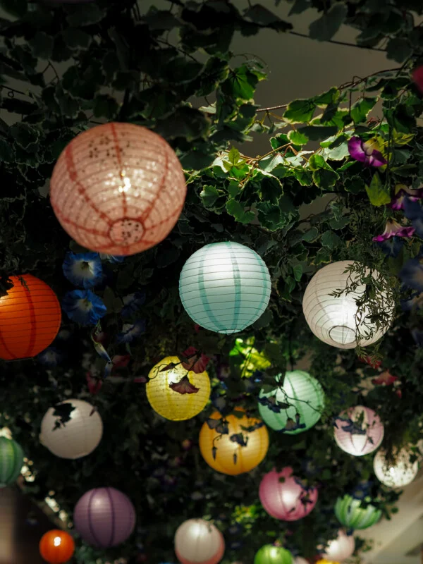 A collection of colorful, glowing paper lanterns hangs from a ceiling covered in lush green foliage and blue flowers.