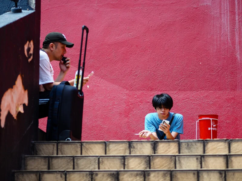 Two men sit on outdoor stairs against a bright red wall, one using a vape and the other holding a smartphone and a cigarette.