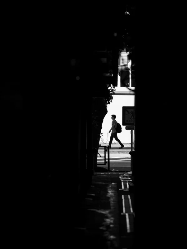 A person with a backpack walks across a bright street as seen through a dark, narrow alleyway.