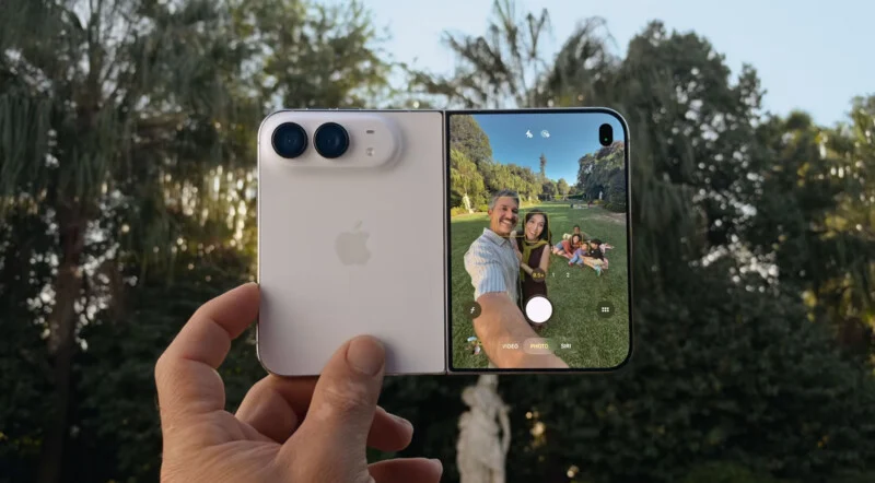 A hand holds a white foldable smartphone, displaying a camera interface on its screen that shows a man and woman taking a selfie in a park.