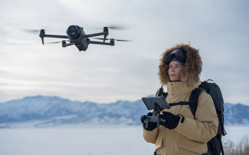 A person in a tan parka and winter gear operates a flying drone with a remote control in a snowy landscape, with mountains visible in the background.