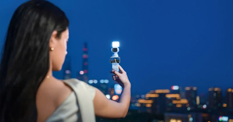A woman seen from behind holds a small, white, illuminated handheld camera toward a blurred city skyline at night.