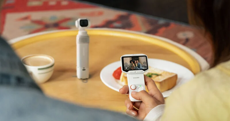 A person holds a white handheld camera with a screen displaying a live feed of three people, while a separate white gimbal camera stands on a wooden table next to a cup of coffee and a plate of food.