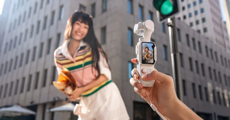 A person holds a small, white, handheld gimbal camera in the foreground, displaying a live view of a smiling woman holding a paper bag on a city street.