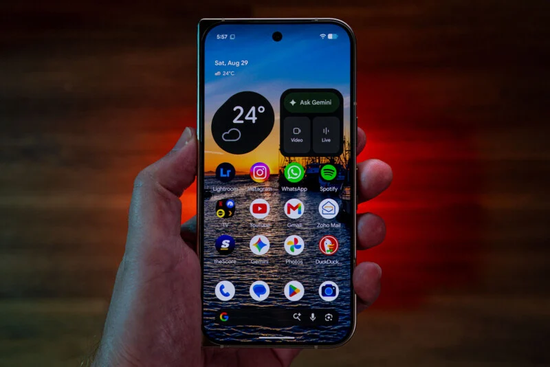 A hand holds a smartphone displaying a home screen with app icons, a weather widget, and a Gemini search bar against a sunset background.