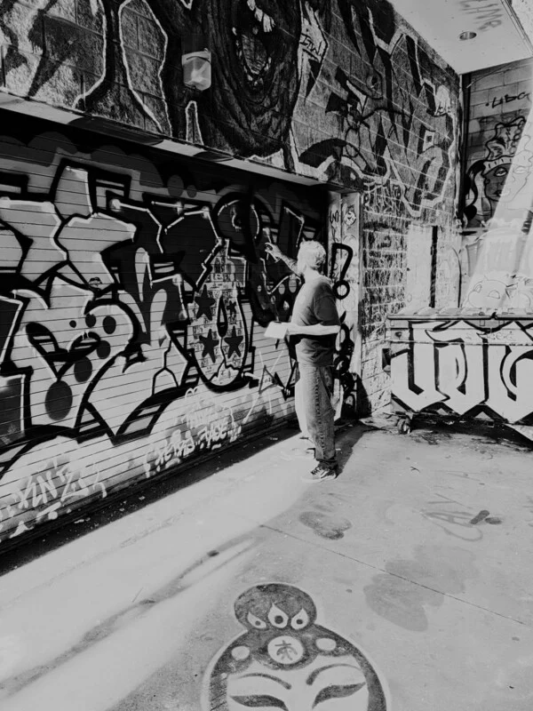 A person stands in an alleyway, spray-painting graffiti onto a metal garage door covered in various artistic designs.