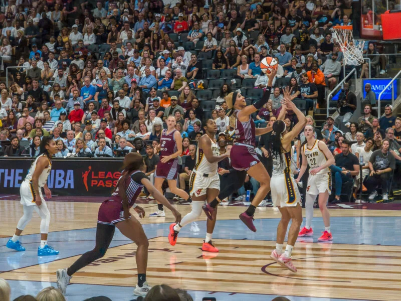 A basketball player in a maroon uniform jumps to shoot a layup while defended by two players in white uniforms during a game.
