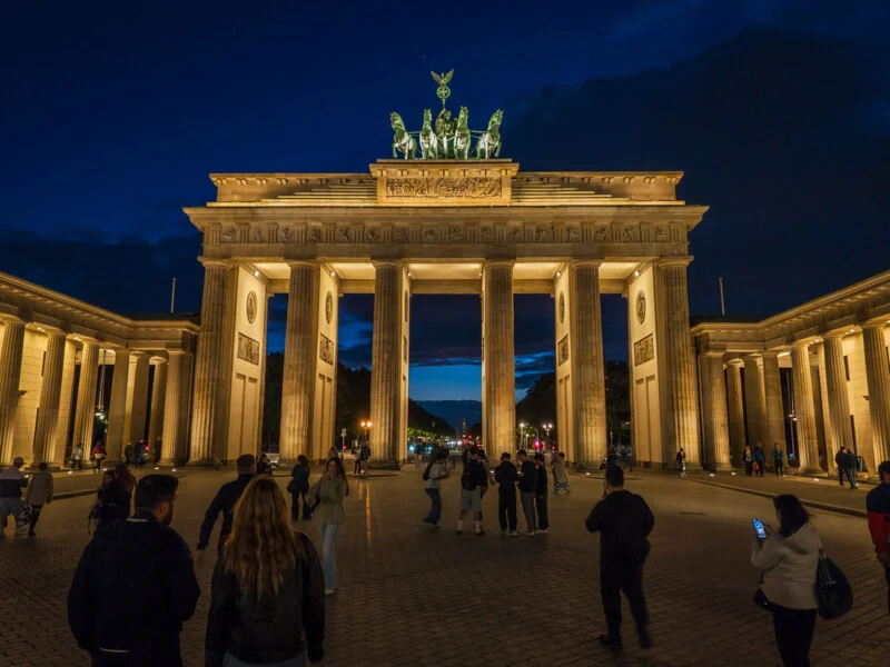 The Brandenburg Gate is illuminated at night, with people walking across the paved square in the foreground.