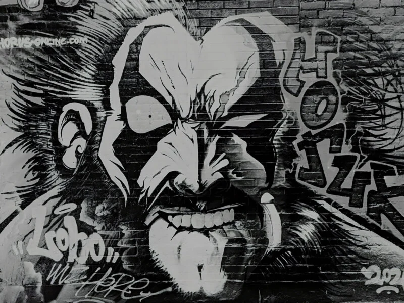 A black and white mural on a brick wall depicts the face of the comic book character Lobo, with graffiti text reading "HORUS-ONLINE.COM" and "Lobo, WE HERE".