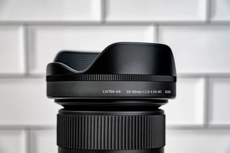 A close-up of a black camera lens hood attached to a lens, with text reading "LH706-04 20-60mm 1:2.8-4 DG ø67 SIGMA" against a white tiled background.
