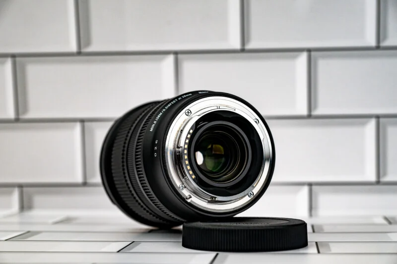 A camera lens rests on its side on a white tiled surface next to a black lens cap, showing its rear mount and electrical contacts.