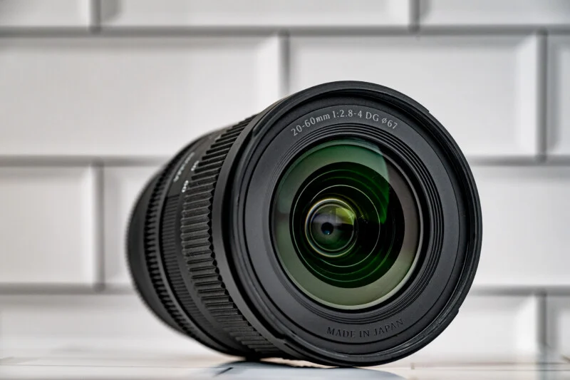 A black camera lens with a 20-60mm 1:2.8-4 DG 67mm specification sits on a white tiled surface against a white brick background.