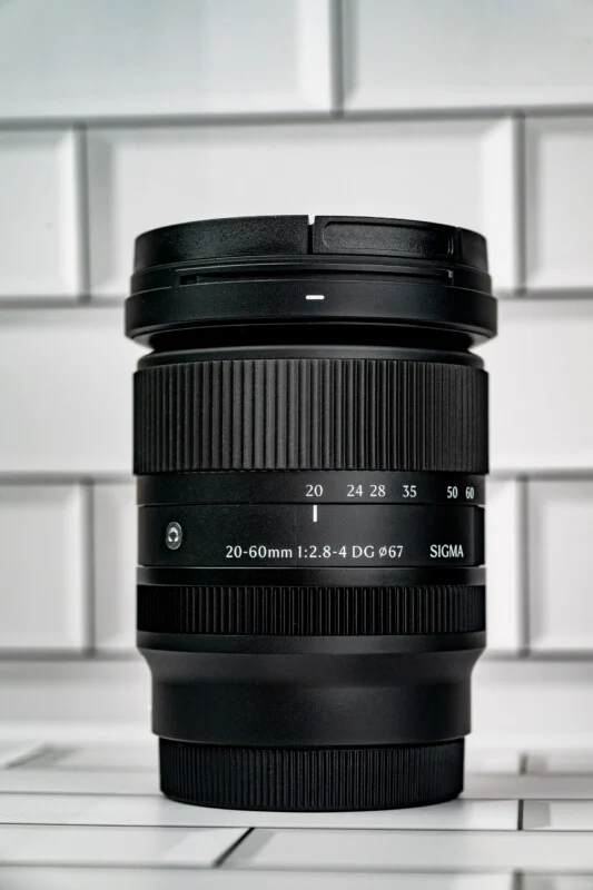A black Sigma 20-60mm 1:2.8-4 DG 67mm camera lens stands upright against a white subway tile background.