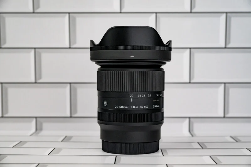 A black Sigma 20-60mm camera lens stands upright against a white subway tile background.