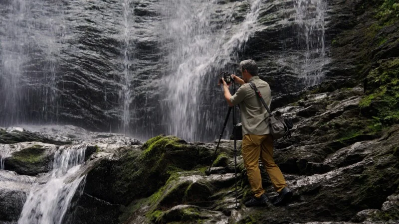 A man with gray hair stands on mossy rocks, using a camera on a tripod to photograph a waterfall.