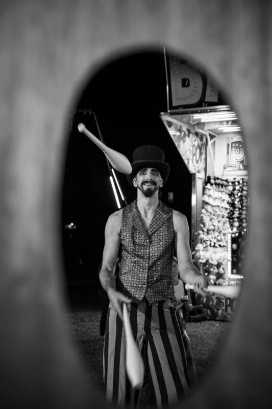 A man in a top hat and patterned vest juggles white clubs while framed by a dark, oval-shaped opening at a carnival.