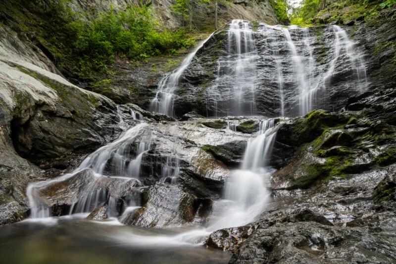 A multi-tiered waterfall cascades over dark, mossy rock formations surrounded by lush green foliage.
