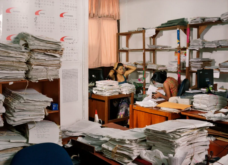 Two women work at desks in an office cluttered with large, stacked piles of paper documents and shelves filled with files.