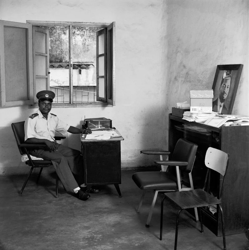 A uniformed man sits at a desk in a sparsely furnished office, with a framed portrait of a man resting on a nearby cabinet.