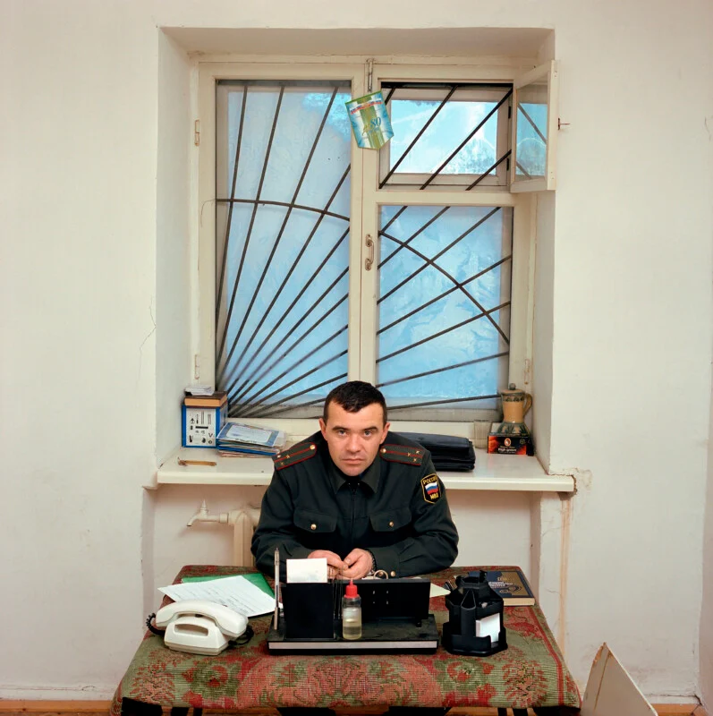 A man in a Russian military uniform sits at a desk in front of a barred window.