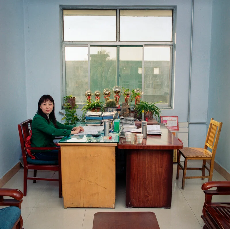 A woman in a green jacket sits at a wooden desk in an office, with trophies and potted plants lined up on the windowsill behind her.
