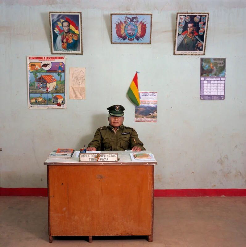 A man in a military uniform sits behind a wooden desk labeled "Director Provincial Tingipaya" in a room decorated with portraits, a flag, and posters.
