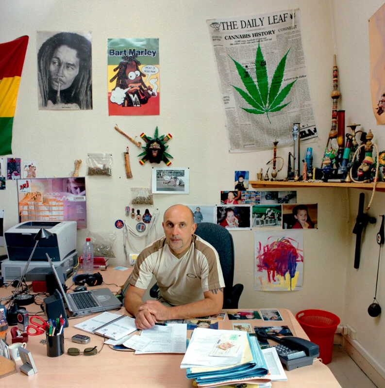 A man sits at a desk in an office decorated with posters of Bob Marley, Bart Simpson, and cannabis-themed imagery.
