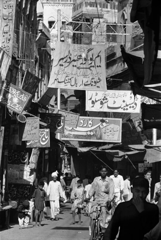 A crowded, narrow street in a South Asian city features numerous hanging signs with Urdu text, pedestrians, and a person riding a bicycle.