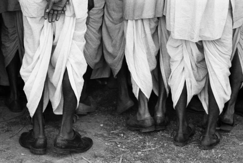 A low-angle, black-and-white shot showing the lower bodies and legs of several men wearing traditional dhotis and dark leather shoes.