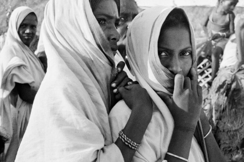 A black-and-white close-up shows two young women wearing headscarves, with one woman's hand resting on the other's shoulder.