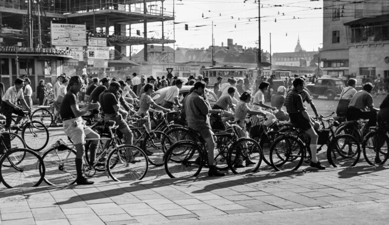 A large group of cyclists waits at a city intersection near a construction site with streetcars and cars in the background.