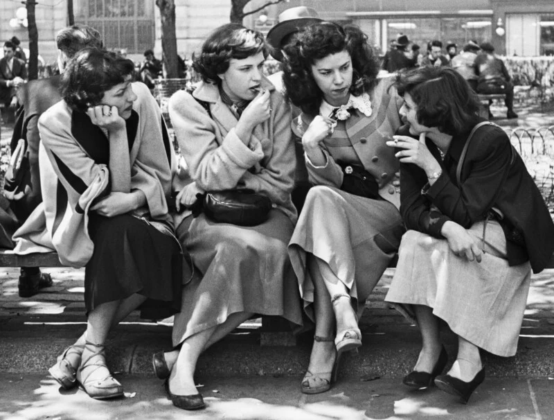 Four women sit together on a park bench, talking and smoking in a black-and-white scene from the mid-20th century.