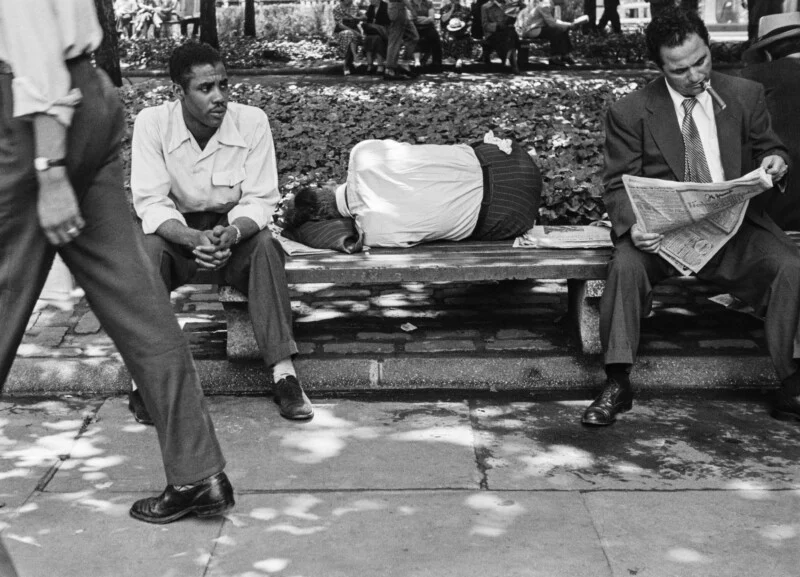 A man sits on a park bench next to someone sleeping, while another man reads a newspaper nearby as a pedestrian walks past in the foreground.